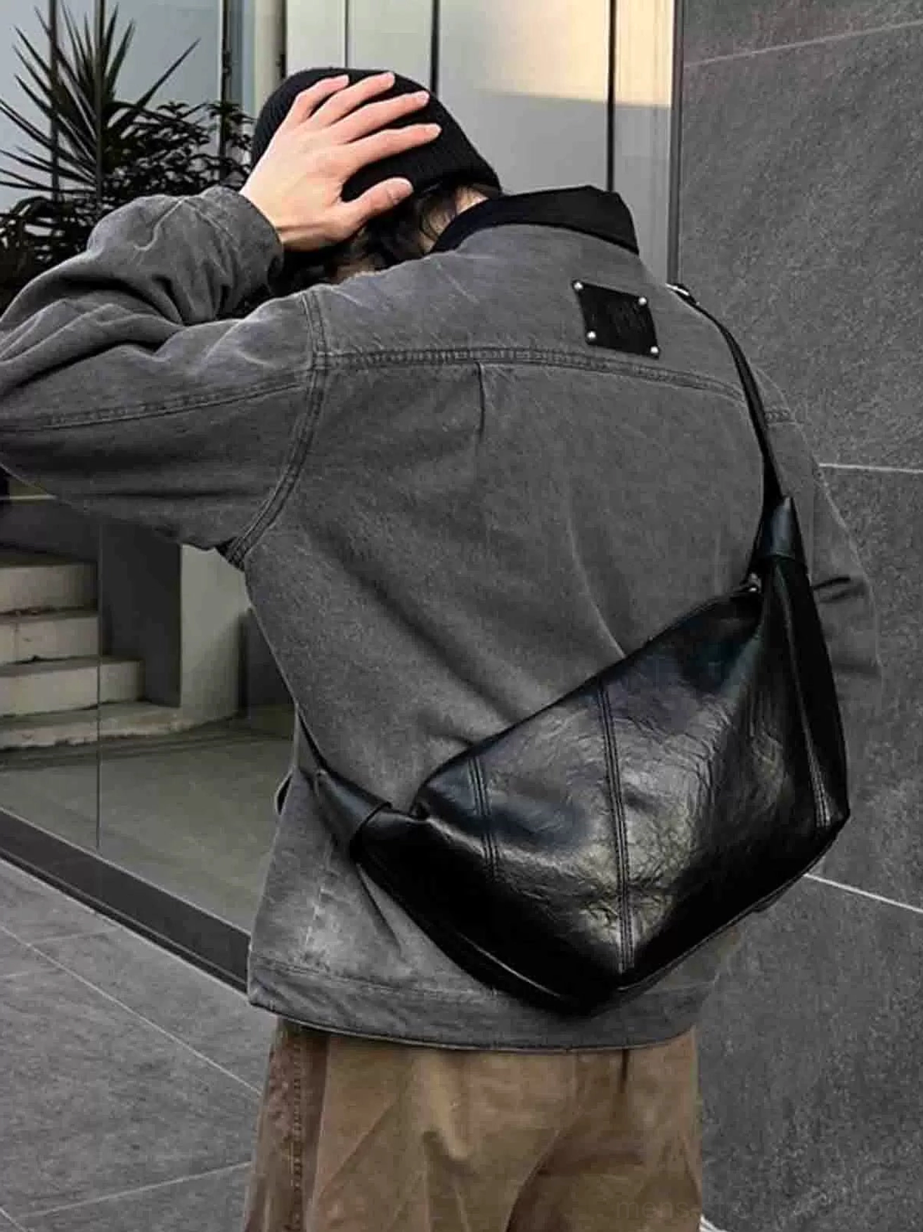 Black minimalist curved PU leather crossbody sling bag worn across the back with gray denim streetwear outfit outdoors