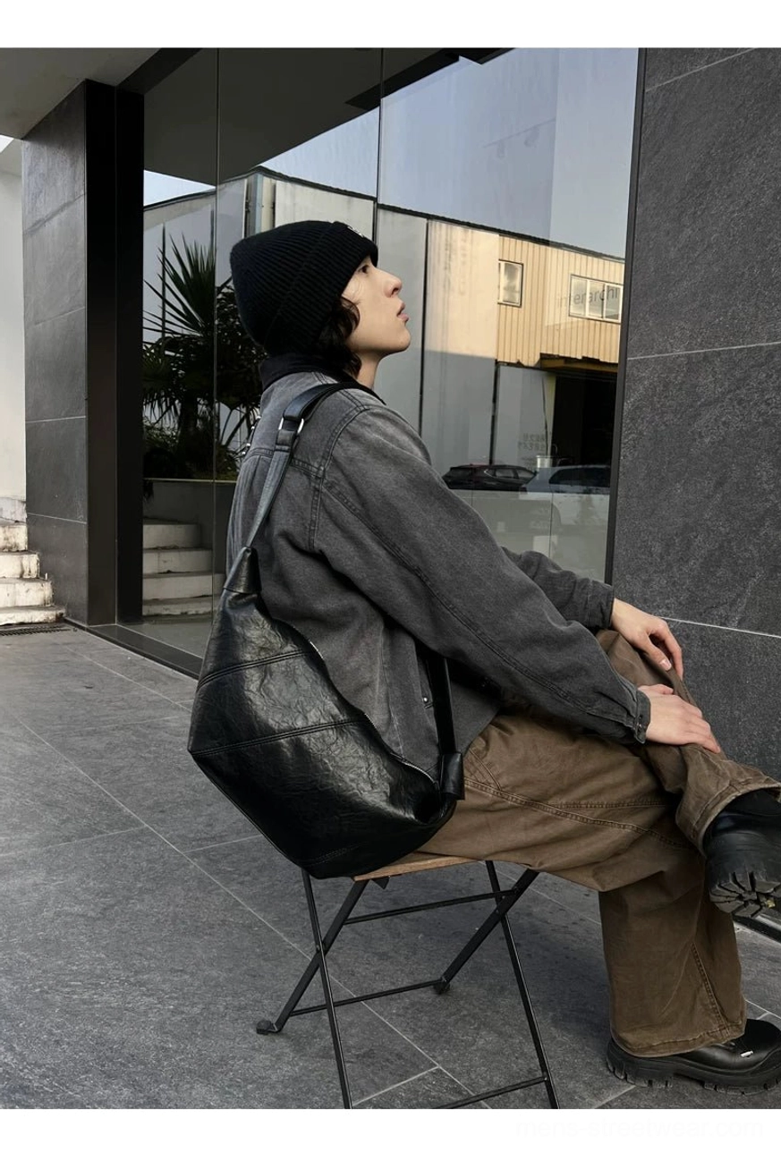 Black minimalist curved crossbody sling bag side view on seated model wearing gray denim urban streetwear