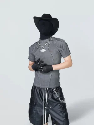 Front full-body view of a dark gray Mirage distressed stitch fitted streetwear tee styled with leather cargo shorts, black cowboy hat, face covering, gloves, and exposed contrast seam details