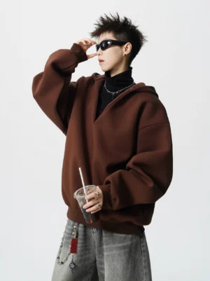 Male model wearing a deep mocha brown oversized V-neck tunnel hoodie with dropped shoulders, black turtleneck, grey wide-leg jeans, and iced coffee in a studio streetwear outfit