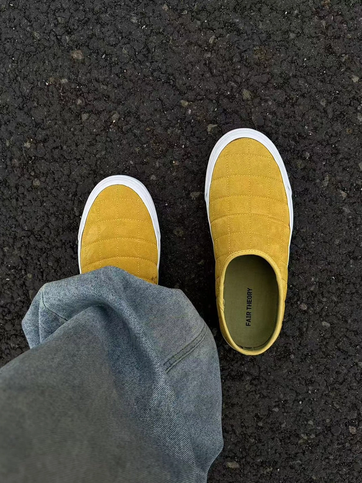 First-person view of mustard yellow quilted slip-on sneaker mules worn with blue jeans on pavement