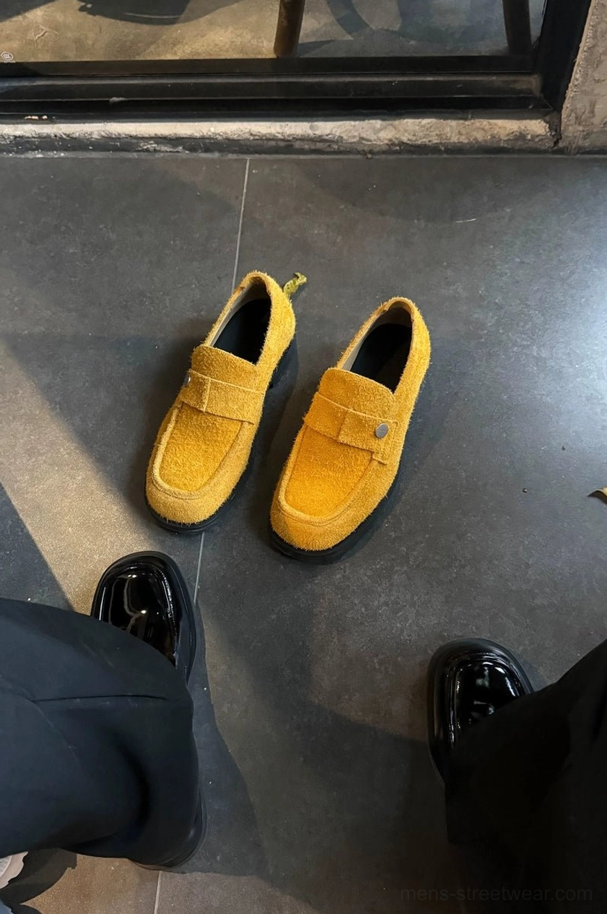 Mustard yellow suede penny loafers worn with black pants outdoors alongside black patent loafers in a contemporary streetwear outfit
