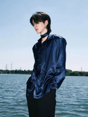 Navy blue satin button-up shirt shown from a side three-quarter view on a male model outdoors beside a lake, featuring a relaxed streetwear fit, glossy fabric, long sleeves, and matching neck tie detail