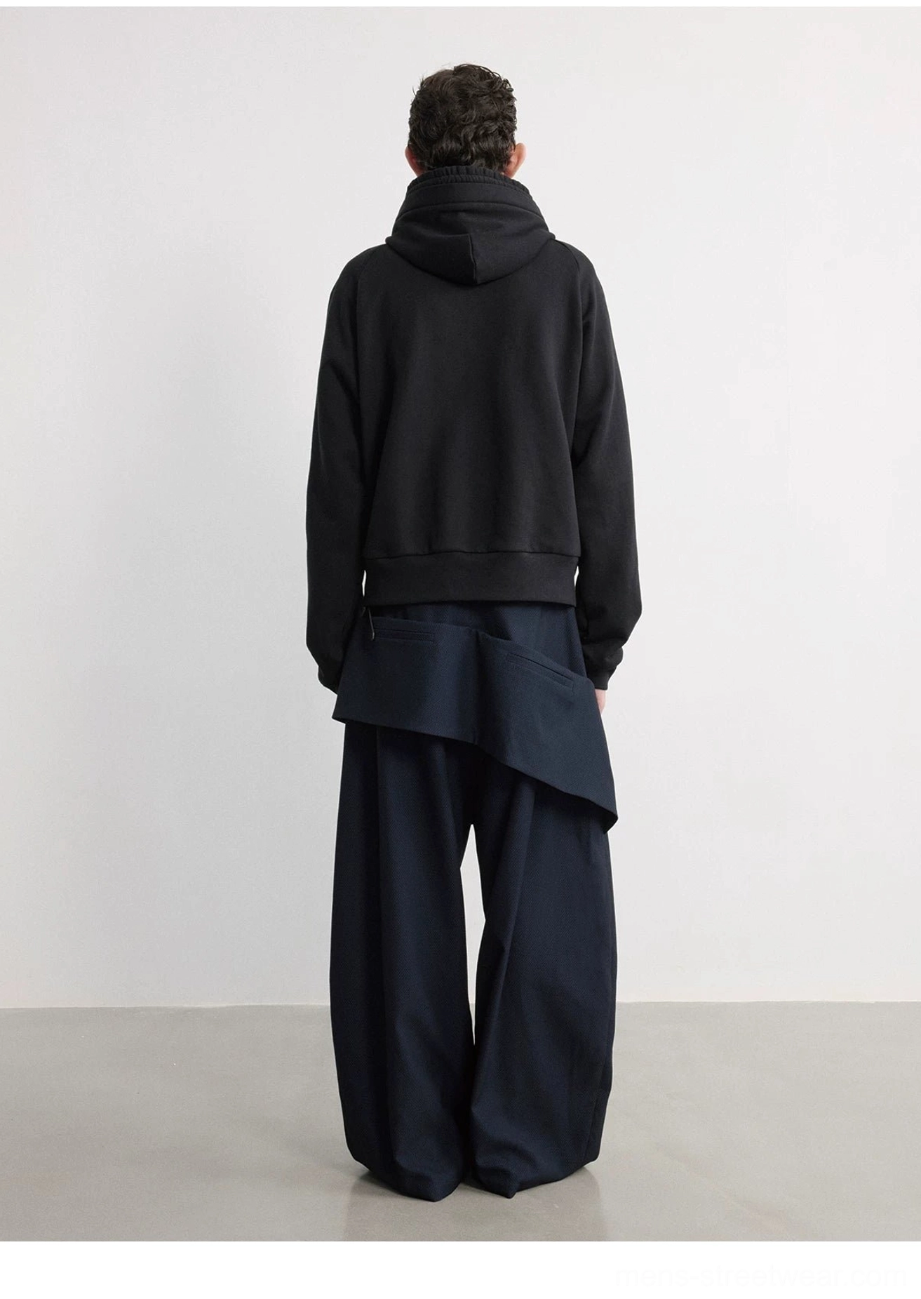 Rear studio view of navy floor-length wide-leg trousers with low-slung asymmetric zipper pouch and black hooded jacket