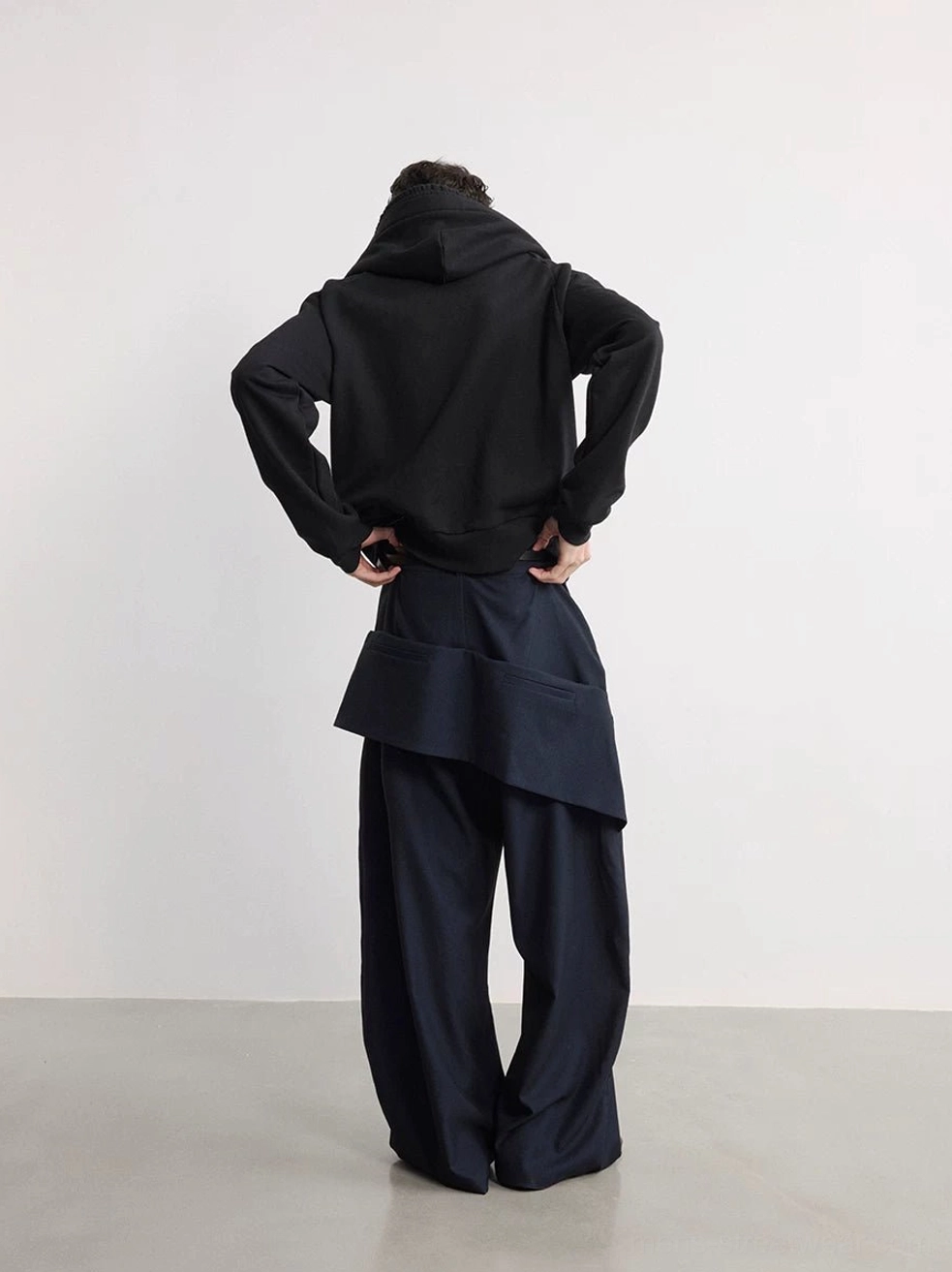 Back view of blue black wide-leg trousers with asymmetric layered zip pouch styled with a black hooded jacket