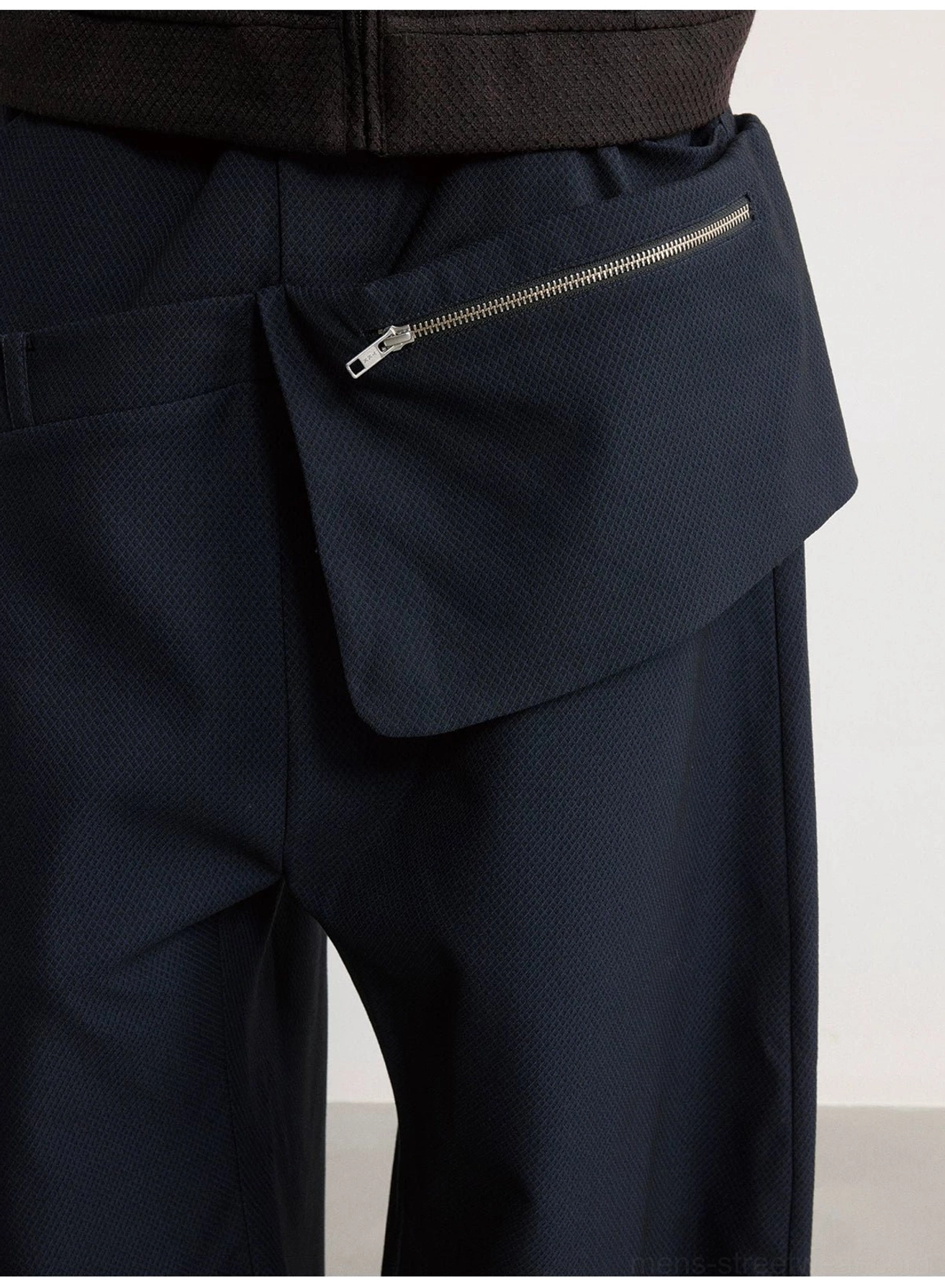 Close-up of textured navy fabric and silver metal zipper on the detachable asymmetric hip pouch of wide-leg trousers
