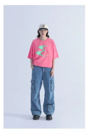 Female model wearing a neon pink oversized contrast logo tee with mint green wave graphic, blue cargo wide leg jeans, and baseball cap