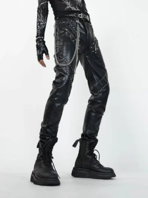 Three-quarter front view of slim black punk leather pants showing cross-shaped thigh rivets, layered silver chains, geometric panels, and fitted streetwear styling