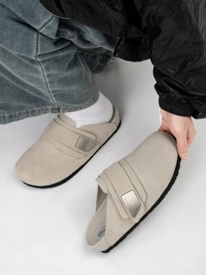 Oatmeal beige suede mule clogs shown from a top-down view with loose denim, featuring an easy slip-on open heel, adjustable strap, and subtle metal detail