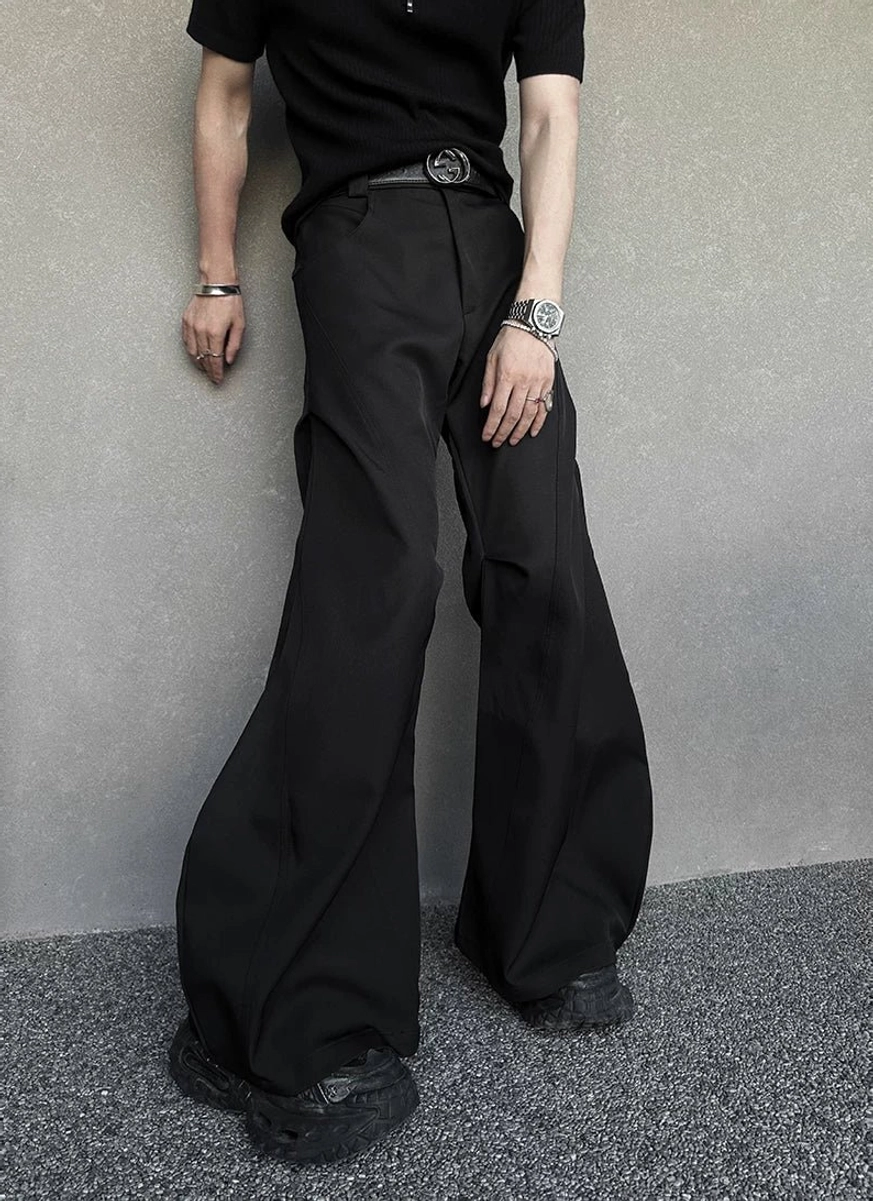 Side view of black hyperwide flare cargo trousers showing vertical leg seam details, round belt buckle, wristwatch, bracelet, and avant-garde streetwear styling