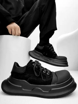 Black chunky platform sneakers worn with all-black streetwear, showing layered suede-finish panels, rounded toe, thick wave-pattern sole, and low-top lace-up design