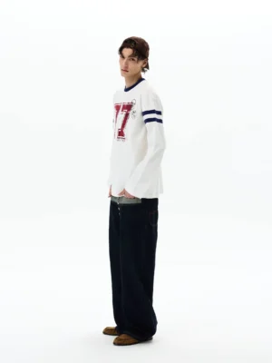 Rear side view of relaxed off-white varsity long sleeve streetwear shirt styled with dark wide-leg jeans and brown loafers