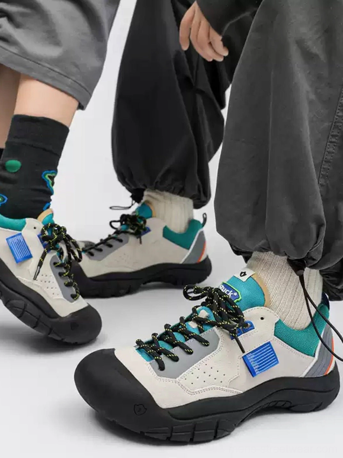 Two models wearing off-white and teal outdoor trail platform sneakers with black rubber toe caps blue accent patches and thick rugged soles in a casual streetwear setting