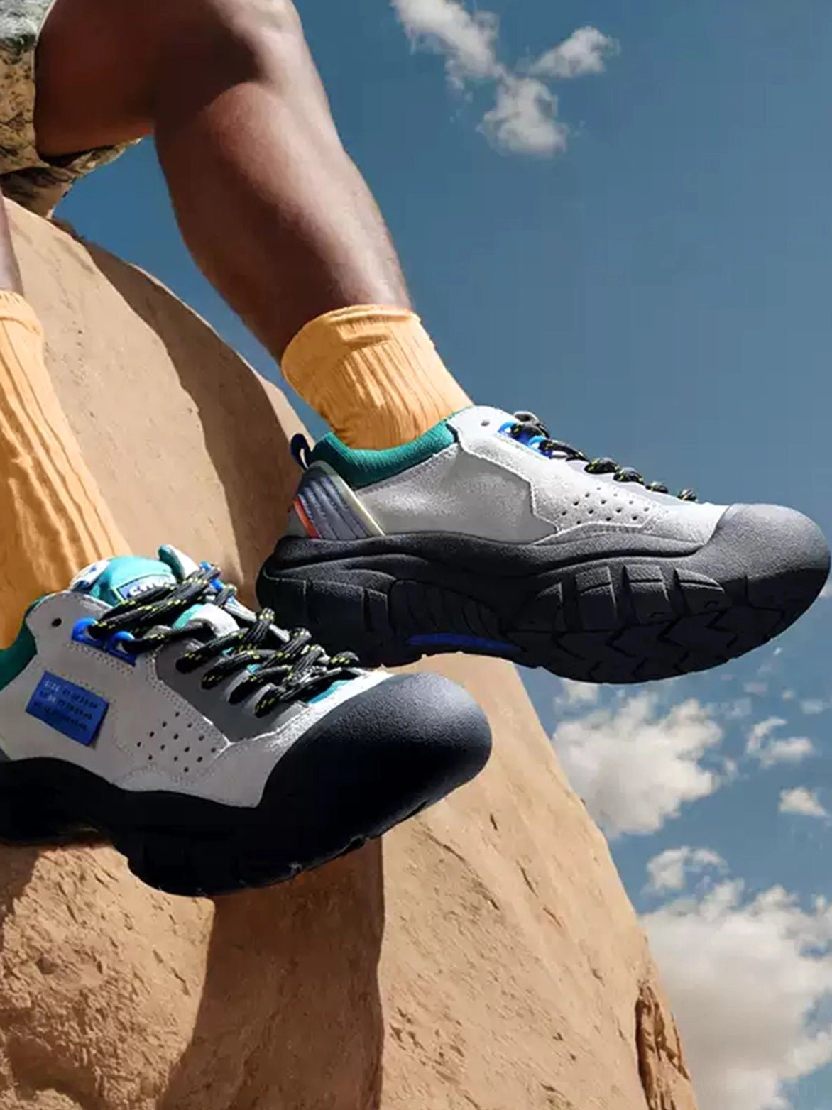 Male model wearing off-white and teal trail platform sneakers with yellow socks on desert rocks under a blue sky, showing the chunky non-slip outsole