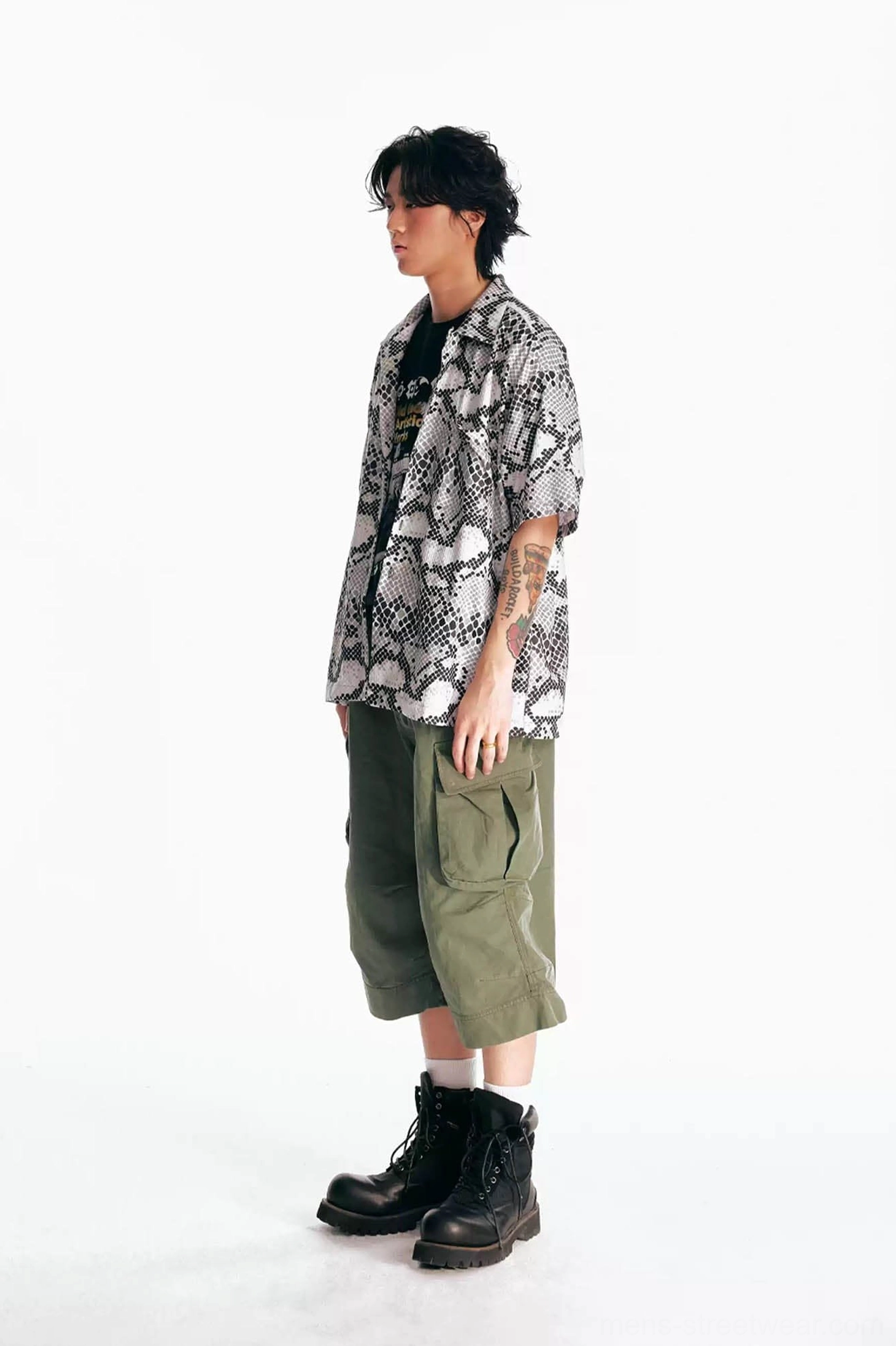 Side view of olive green flared utility cargo Bermuda shorts styled with a paisley short sleeve shirt, black inner top, and black work boots