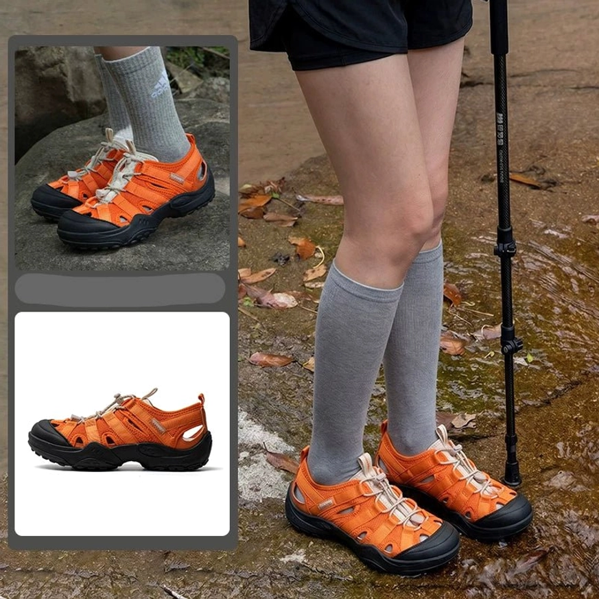 Bright orange outdoor water hiking sandals worn with gray socks on a leafy rocky hiking path, showing black trim, bungee lacing, and durable rubber sole