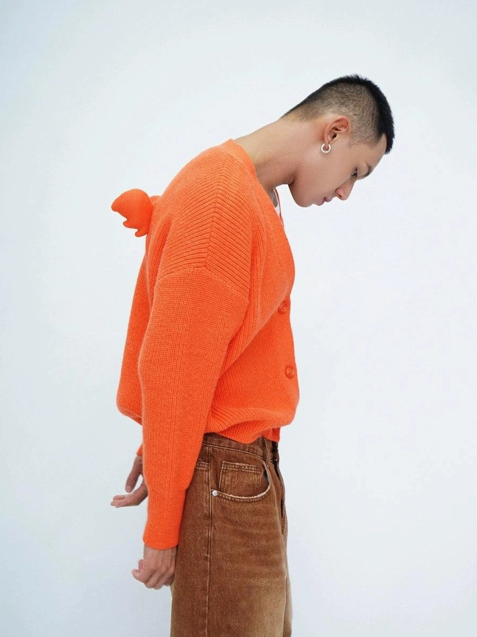 Male model wearing a bright orange oversized fuzzy knit cardigan with ribbed texture and wing-shaped pom-pom back detail