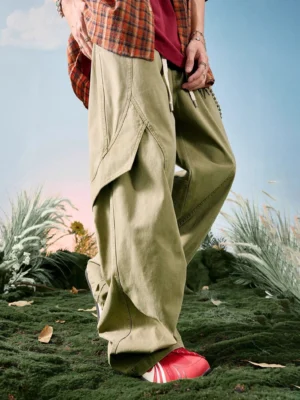 Khaki relaxed wide leg cargo pants shown from a low rear side angle with oversized utility pocket, adjustable drawstring waistband, decorative metal chain, and red stripe sneakers in an outdoor streetwear setting