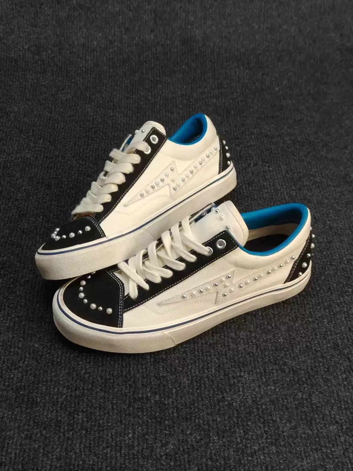 Overlapping low-top skate sneakers with white paneled uppers, black toe sections, bright blue inner collars, and pearl-like studs along the vamp and heel