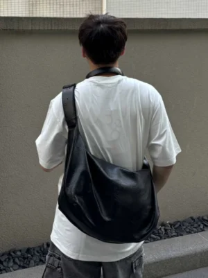 Black oversized crescent sling bag worn crossbody with a relaxed draped silhouette on a male streetwear model in an urban setting