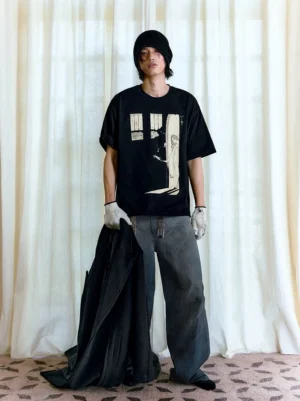 Male model wearing an oversized black gothic graphic T-shirt with cathedral window and doorway artwork, styled with a beanie, gloves, baggy jeans, and a black coat
