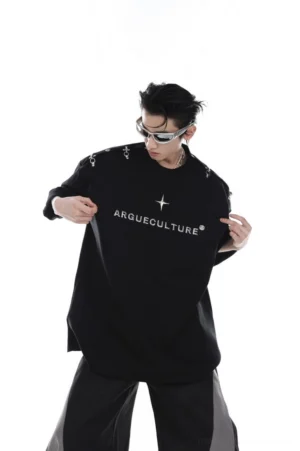 Front full-body view of an oversized black metallic logo T-shirt with silver chest lettering, star metal accent, and shoulder buckle details styled with futuristic sunglasses and wide-leg pants