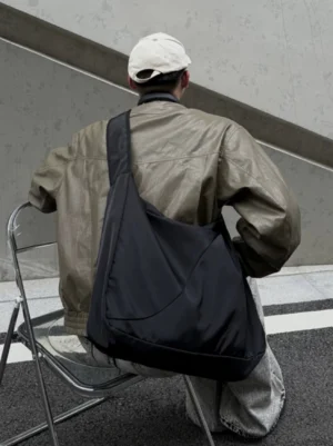 Oversized black nylon city sling bag worn across the back with khaki jacket in an industrial streetwear setting
