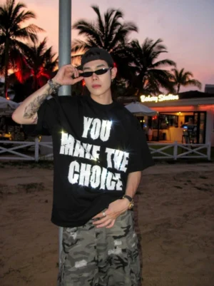 Front view of oversized black streetwear graphic tee with sparkling YOU MAKE THE CHOICE lettering, styled with camouflage cargo pants at a seaside sunset