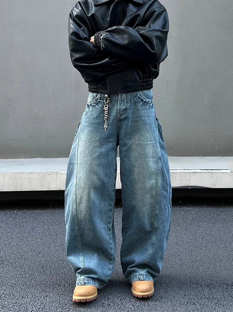 Front full-body view of oversized blue balloon fit jeans with curved side seams, vintage faded denim wash, cropped black leather jacket, metal waist chain, and tan work boots in an urban streetwear setting