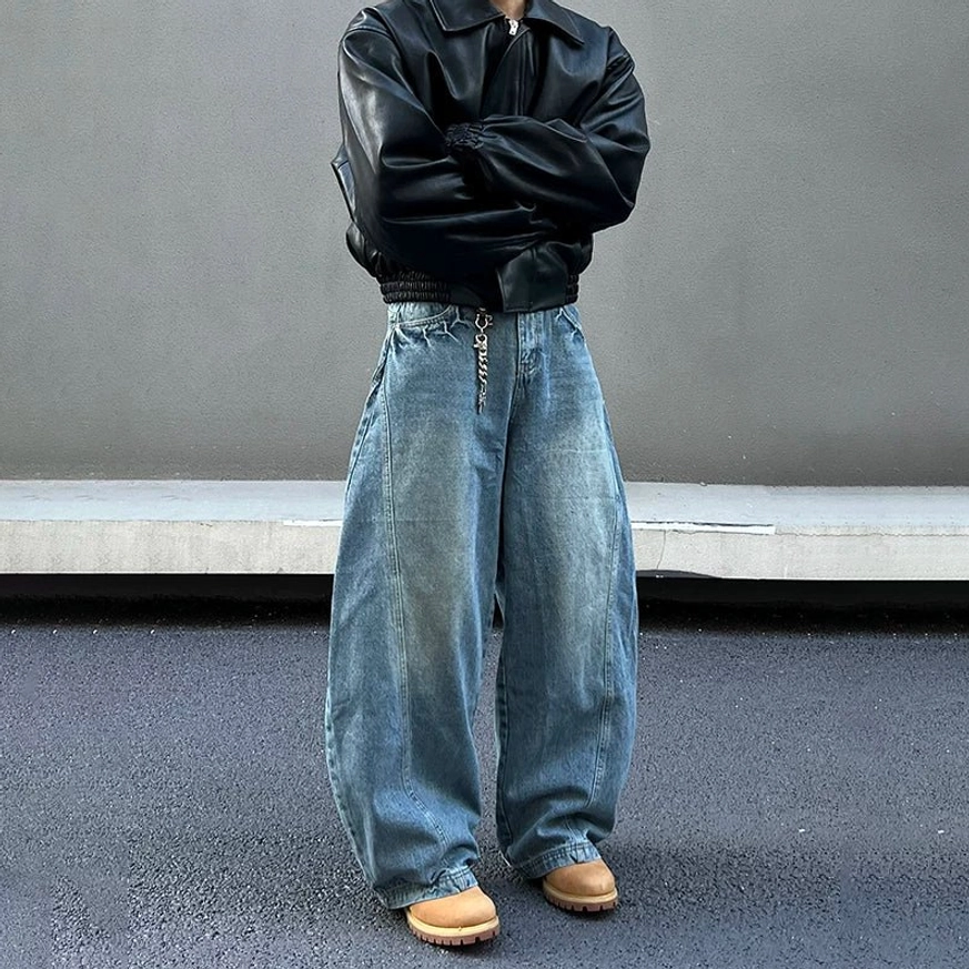 Three-quarter front view of oversized blue balloon jeans styled with a cropped black leather jacket, metal waist chain, tan work boots, and urban Korean streetwear attitude
