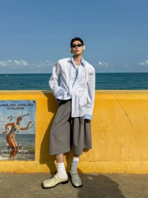 Oversized blue and white colorblock stripe button-up shirt worn by male model at seaside yellow guardrail front view