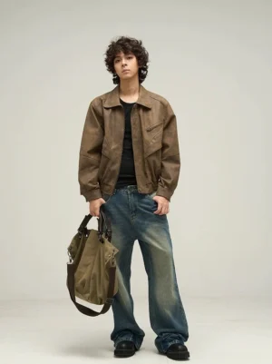 Full-body view of a man wearing an oversized brown faux leather jacket with wide-leg blue jeans, black shoes, and a khaki shoulder bag