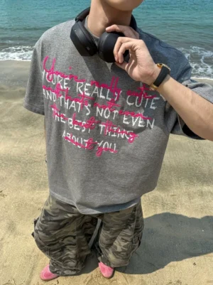 Close-up front view of an oversized charcoal grey graffiti slogan T-shirt with pink and white handwritten compliment graphic, styled with camouflage wide-leg pants, pink socks, and black headphones