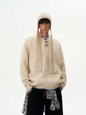 Apricot oversized contrast-stitch Henley knit sweater shown on a male model with a relaxed Korean streetwear silhouette, layered plaid shirt detail, and matching knitted headwear