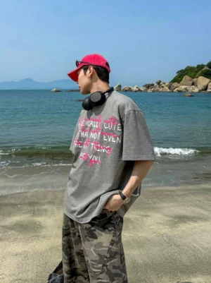 Side profile of a male model wearing an oversized dark grey graphic streetwear T-shirt with pink and white graffiti lettering, red baseball cap, and casual beach styling