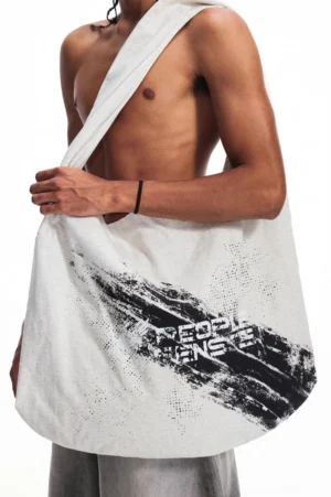 Oversized distressed gray canvas tote bag with black brushstroke graphic print worn over one shoulder in a men's streetwear outfit
