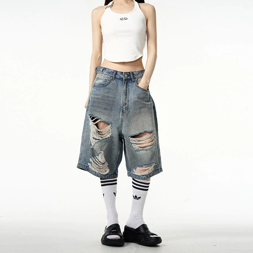 Front full-body streetwear styling of oversized distressed ripped denim Bermuda shorts with a white cropped tank top, striped socks, and black loafers
