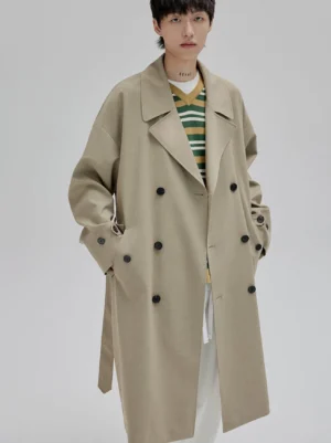 Tan oversized double-breasted long trench coat shown from the front three-quarter view with wide lapels, black buttons, adjustable waist belt, and relaxed dropped shoulders