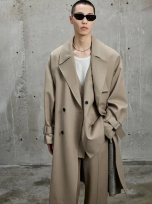 Khaki oversized double-breasted long trench coat shown from the front with wide notched lapels and relaxed dropped shoulders against an industrial concrete wall