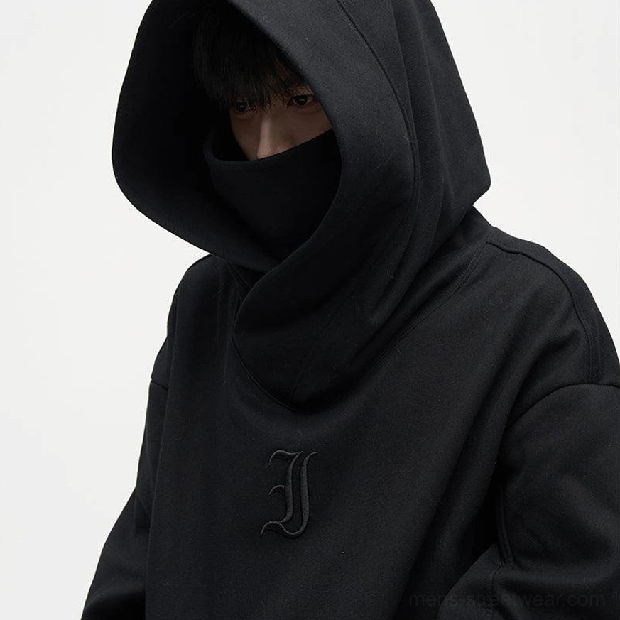 Black oversized dual-layer high-neck hoodie close-up showing face-covering hood construction and tonal embroidered chest detail
