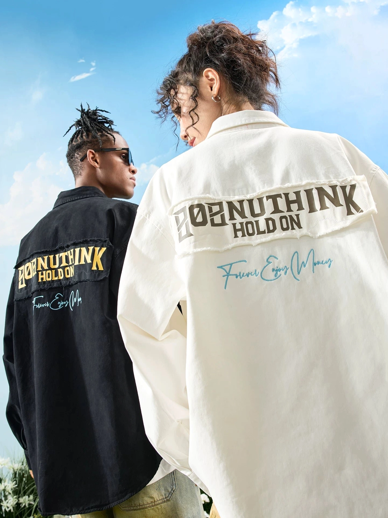 Two models wearing black and off-white oversized embroidered button-up shirts with bold back graphics in a sunny outdoor streetwear setting