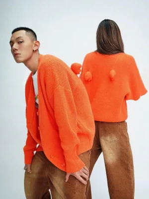 Bright orange oversized fuzzy knit cardigan shown from front and back with ribbed texture, wing-shaped pom-pom back details, and wide-leg streetwear styling