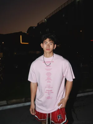 Pink oversized graphic streetwear t-shirt with vertical typography print styled with red basketball shorts and silver necklace on a nighttime city street