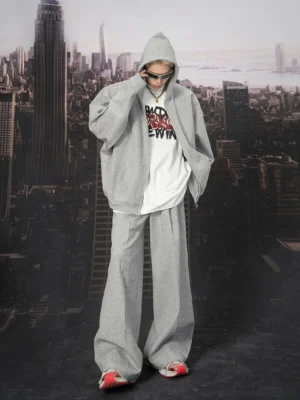 Male model wearing oversized heather grey zip-up hoodie with hood up, layered white graphic tee and casual sneakers