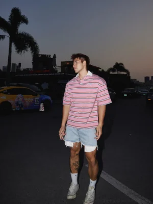 Side view of loose fit pink striped short sleeve rugby polo styled with distressed denim shorts and sneakers