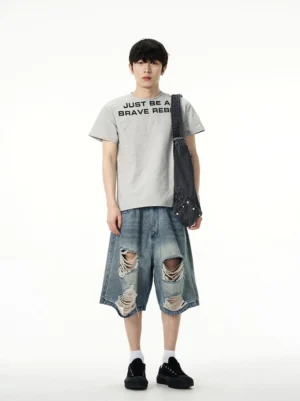 Male model wearing oversized ripped denim Bermuda shorts with a grey graphic T-shirt and black crossbody bag in a relaxed urban streetwear outfit