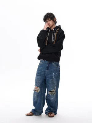 Black oversized tri-cord drawstring hoodie shown from the side with dropped shoulders, relaxed sleeves, and contrasting hood cords styled with distressed streetwear denim