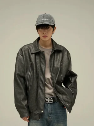 Black oversized vintage faux leather streetwear jacket shown from the front with a relaxed drop shoulder fit, spread collar, zipper pockets, beige tee, blue jeans, and baseball cap