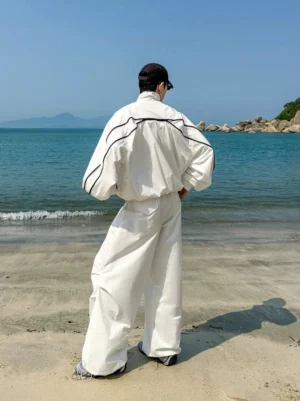 Back three-quarter view of oversized white streetwear zip bomber jacket showing black contrast back yoke and shoulder piping by the sea