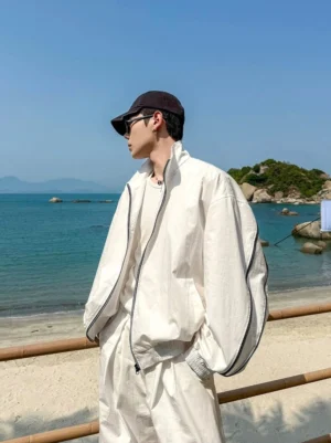 Oversized white contrast piping zip bomber jacket worn in side profile on a coastal beach with stand collar and black sleeve trim